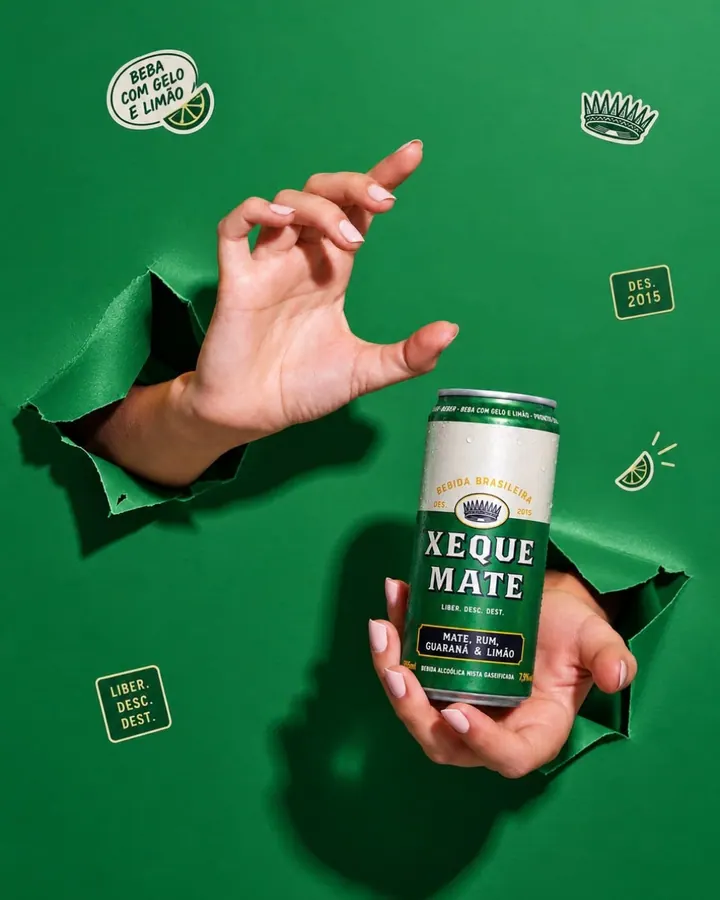 Mãos atravessando um papel verde rasgado e segurando a lata Xeque Mate, com adesivos da marca em volta
